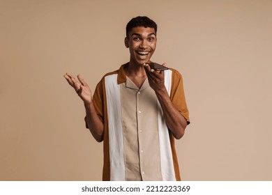 Young African Handsome Enthusiastic Man Recording Audio Message On Phone And Looking At Camera , While Standing Over Isolated Beige Background