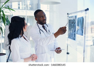 Young african male and caucasian female doctors discussing MRI results of patient in the hospital. Male and female doctors in white coats with stethoscopes. Medical and health care concept.