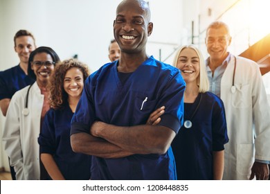 Young African male doctor smiling while standing in a hospital corridor with a diverse group of staff in the background - Powered by Shutterstock