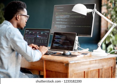 Young african male programmer writing program code sitting at the workplace with three monitors in the office. Image focused on the screen