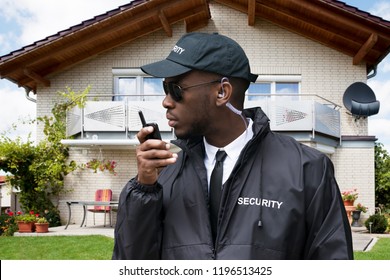 Young African Male Security Guard Talking On Walkie Talkie In Front Of House