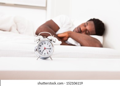Young African Male Sleeping On Bed In Front Of Alarm Clock