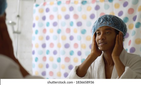 Young African Man In Bathrobe Looking Into Mirror Putting On Shower Cap Before Taking Bath. Reflection Of Cheerful Afro-american Guy In Shower Cap Standing In Bathroom In Morning