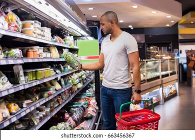 Young African Man Buying Vegetables In Grocery Section At Supermarket. Black Man Choose Vegetables In The Supermarket While Holding Grocery Basket. Man Shopping Veggies At Supermarket.