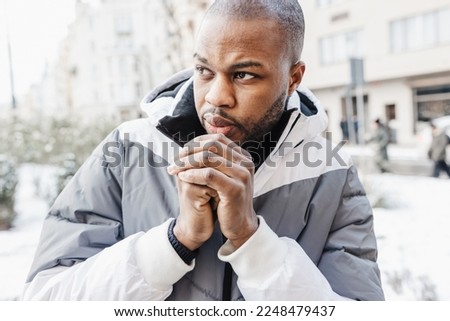 Young African man in casual white and blue clothes, warming his cold hands with breath. Winter season street portrait with snow. Lifestyle, city life concept. Close up