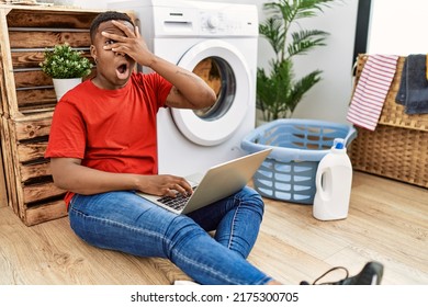 Young African Man Doing Laundry And Using Computer Peeking In Shock Covering Face And Eyes With Hand, Looking Through Fingers With Embarrassed Expression. 