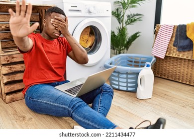 Young African Man Doing Laundry And Using Computer Covering Eyes With Hands And Doing Stop Gesture With Sad And Fear Expression. Embarrassed And Negative Concept. 