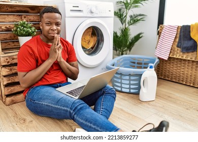 Young African Man Doing Laundry And Using Computer Praying With Hands Together Asking For Forgiveness Smiling Confident. 