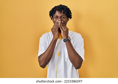 Young African Man With Dreadlocks Standing Over Yellow Background Laughing And Embarrassed Giggle Covering Mouth With Hands, Gossip And Scandal Concept 