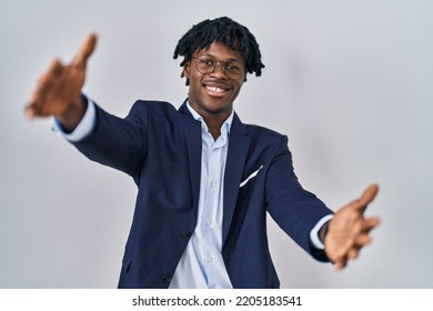 Young African Man With Dreadlocks Wearing Business Jacket Over White Background Looking At The Camera Smiling With Open Arms For Hug. Cheerful Expression Embracing Happiness. 