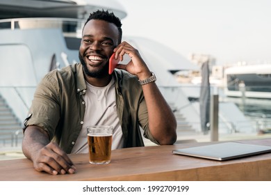 Young african man drinking beer at brewery bar using mobile phone with luxury yacht port in background - Powered by Shutterstock