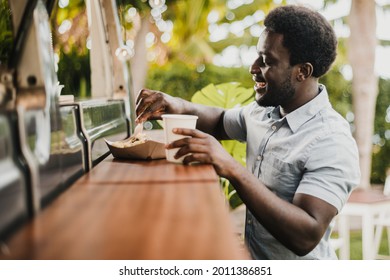 Young African Man Eating In Food Truck Counter Outdoor In City Park - Focus On Face