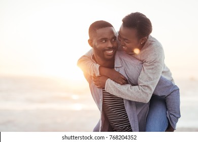 Young African man giving his laughing girlfriend a piggyback while enjoying a late afternoon together at the beach