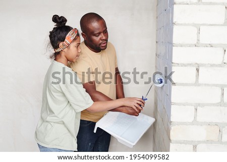 Young African man helping his cute daughter with paintroller painting brick wall of living-room into white color while standing in the corner