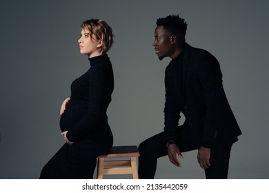 Young African Man And His Caucasian Pregnant Woman Sit Against Grey Background. Interracial Marriage Concept.