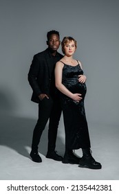 Young African Man And His Caucasian Pregnant Woman Stand Against Grey Background. Interracial Marriage Concept.