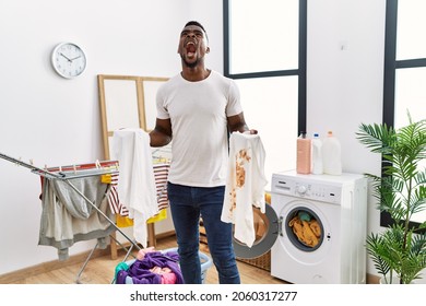 Young African Man Holding Clean White T Shirt And T Shirt With Dirty Stain Angry And Mad Screaming Frustrated And Furious, Shouting With Anger Looking Up. 