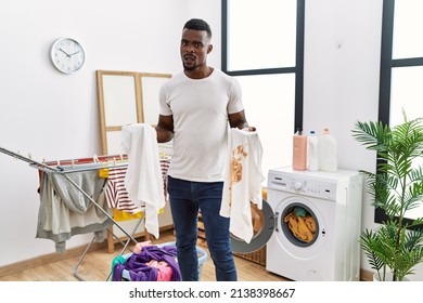 Young African Man Holding Clean White T Shirt And T Shirt With Dirty Stain Clueless And Confused Expression. Doubt Concept. 