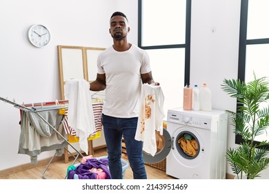 Young African Man Holding Clean White T Shirt And T Shirt With Dirty Stain Puffing Cheeks With Funny Face. Mouth Inflated With Air, Catching Air. 