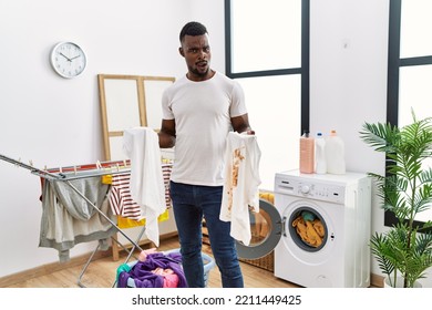 Young African Man Holding Clean White T Shirt And T Shirt With Dirty Stain In Shock Face, Looking Skeptical And Sarcastic, Surprised With Open Mouth 