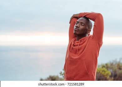 Young African man in a hoodie standing on a trail by the ocean stretching before a cross country run - Powered by Shutterstock