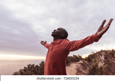 Young African Man In A Hoodie Standing With His Eyes Closed And Arms In The Air Embracing Nature, While Out For A Run By The Ocean