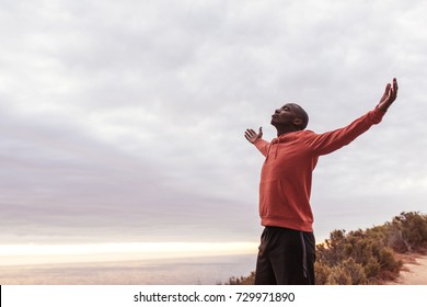 Young African man in a hoodie standing with his arms raised to the sky while out for a cross country run by the ocean - Powered by Shutterstock