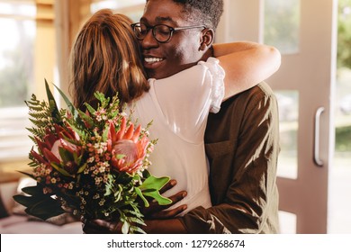 Young African Man Hugging His Girlfriend Standing In Cafe. Young Guy Expressing His Love For His Lady Giving Flowers And A Warm Hug During A Coffee Shop Date.