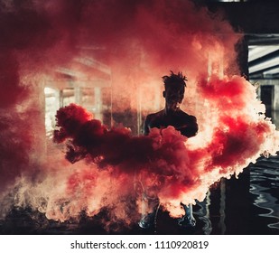 Young African Man In Jeans Stands In Water Under Bridge And Holds Colored Red Smoke Bomb In His Hands On Background Of Concrete Supports. With Backlight.