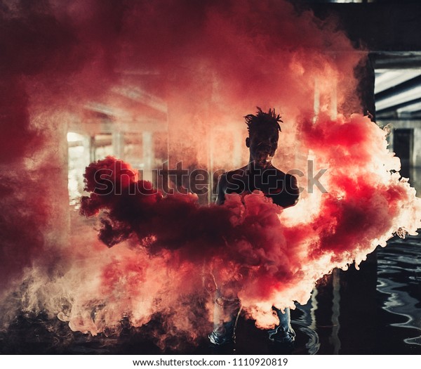 Young African man in jeans stands in
water under bridge and holds colored red smoke bomb in his hands on
background of concrete supports. With
backlight.