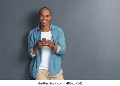 Young african man leaning against a grey wall using mobile phone. Portrait of a happy laughing guy texting a phone message and looking at camera. Man reading a message on cell phone with copyspace. - Powered by Shutterstock