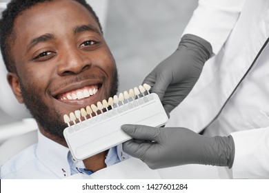 Young African Man Looking At Camera And Smiling While Female Dentist Keeping Range Of Fillings In Clinic. Happy Patient Enjoying White Beautiful Smile While Visiting Dentist Office. Concept Of Cure.