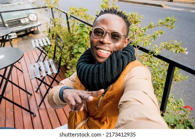 Young African Man Outdoors, Sitting Outside Cafe Happy Smiling Takes Selfie Making V Sign With Hand, Looking At Camera, Perspective From Phone.