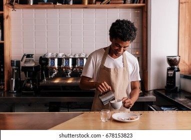 Young african man pouring milk into coffee making espresso. Professional barista preparing coffee on counter. - Shutterstock ID 678153823