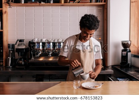 Young african man pouring milk into coffee making espresso. Professional barista preparing coffee on counter.