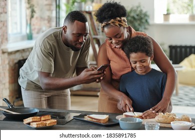 Young African man with smartphone taking photo of his cute little son cutting bread for sandwiches while preparing breakfast for family - Powered by Shutterstock