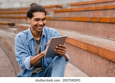 Young African Man Student Freelancer Using Digital Tablet Reading E-book, Working Remotely, Surfing On Internet, Social Media, Checking E-mails, E-learning, E-banking Online In Urban Area