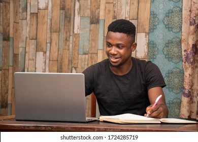 young african man studying at home using his laptop, receiving lectures online and taking notes, smiling