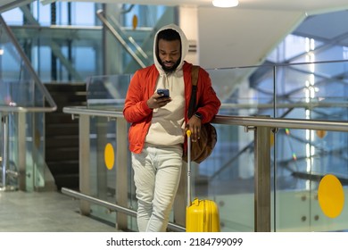 Young African Man Traveler With Baggage Booking Taxi Via Mobile App, Black Guy Surfing Internet Or Connecting To Free Wifi While Waiting For Flight In Modern Airport Terminal. Tourism And Technology