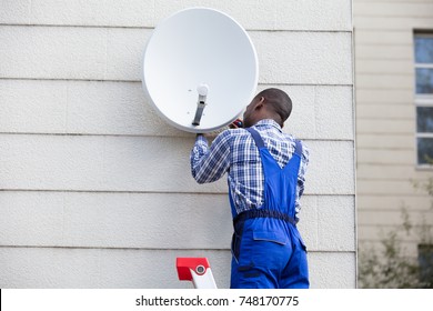 Young African Man In Uniform Fitting TV Satellite Dish On Wall