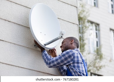 Young African Man In Uniform Fitting TV Satellite Dish On Wall