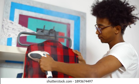 Young African Man Using Steamer On Checkered Shirt Before Getting Dressed. Afro-american Guy In Glasses Steaming Clothes On Rack At Home.