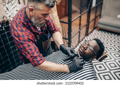 Young African man visiting hairstylist in barber shop. Professional hairdresser shaving beard with electric shearer machine.  - Powered by Shutterstock