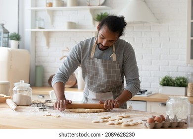 Young African man wear apron rolls out dough for cookies standing alone in kitchen, holds rolling-pin flattening dough make pastry, homemade biscuits, prepare sweets for family. Cookery, hobby concept - Shutterstock ID 2089628230