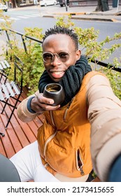 Young African Man Wearing Glasses, Scarf And Winter Clothes, Makes Selfie With His Phone Drinking Coffee Sitting Outside Coffee Shop, Looking At Camera, Phone Perspective.