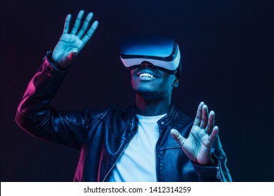 Young african man wearing virtual reality goggles with hands up, isolated on black background