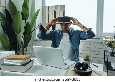 Young African Man Wearing Virtual Reality Glasses While Sitting At Working Place               