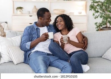 Young African Man And Woman Enjoying Weekend Together, Sitting On Couch Together At Home, Drinking Tea And Talking