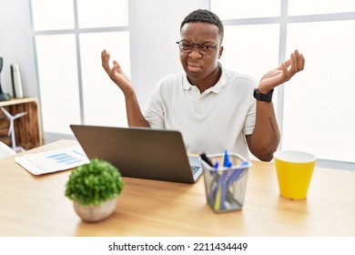 Young African Man Working At The Office Using Computer Laptop Clueless And Confused Expression With Arms And Hands Raised. Doubt Concept. 