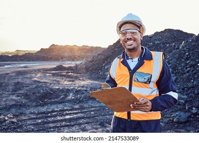A young African mine worker wearing protective wear is looking at the camera while holding a clipboard with coal mine in the background - Shutterstock ID 2147533089